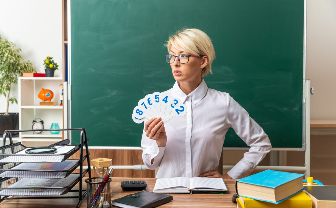serious young blonde female teacher wearing glasses sitting at desk with school tools in classroom holding number fans keeping hand on waist looking at side