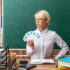 serious young blonde female teacher wearing glasses sitting at desk with school tools in classroom holding number fans keeping hand on waist looking at side