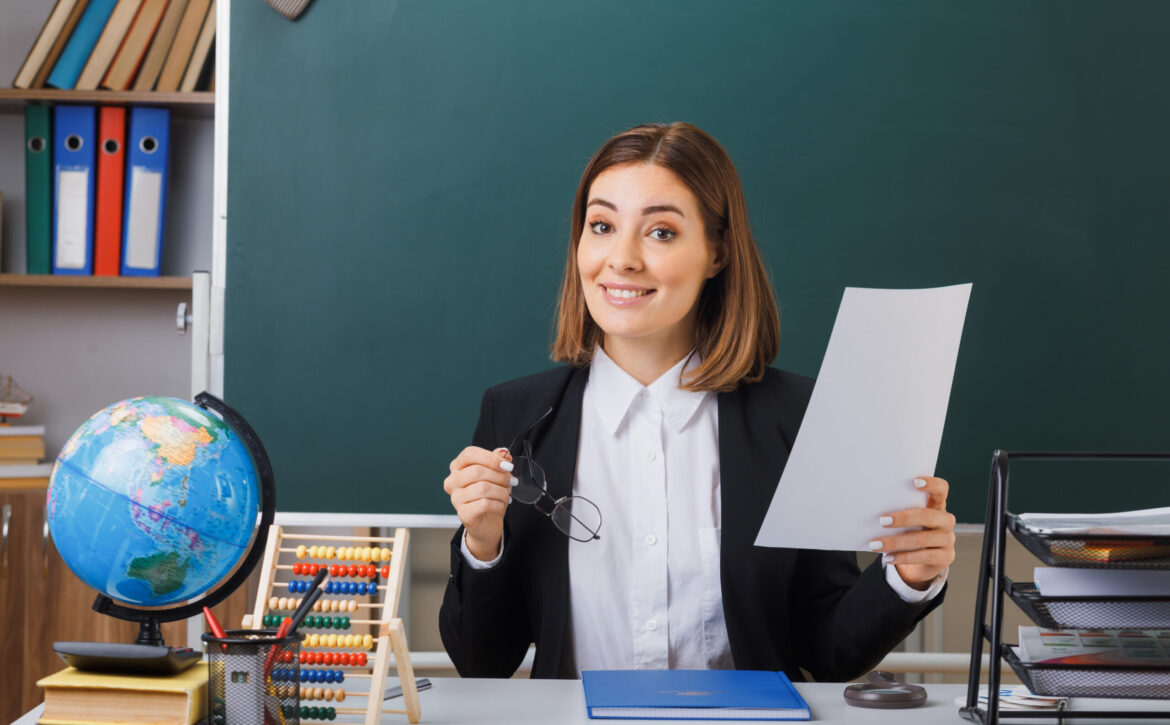 young woman teacher wearing glasses sitting at school desk with globe and books in front of blackboard in classroom holding white empty sheet of paper happy and positive smiling